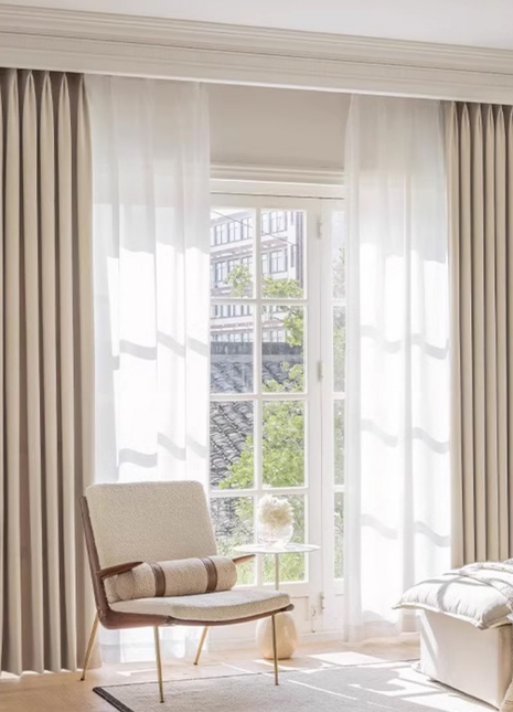 a well-lit living room with a comfortable chair, a glass table, and large windows with sheer curtains.
