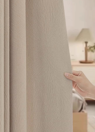 A hand is holding a beige curtain, which is partially open, revealing a lamp and a wooden dresser in the background.