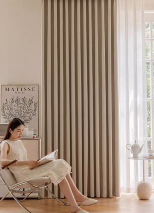 A woman in a white dress is sitting in a chair and reading a book in a room with a large window and a beige curtain.