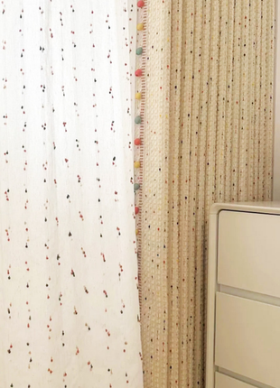 a white curtain with multicolored dots and a beige curtain with a similar pattern. The curtains are hanging next to a white dresser.