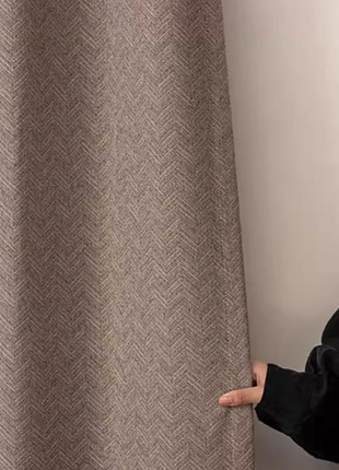 A person is holding a brown and beige patterned curtain in front of a white wall.