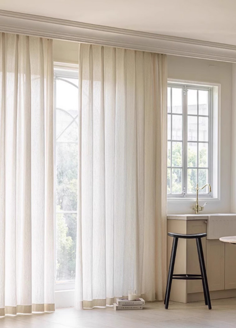 a bright and airy kitchen with a large window, white curtains, and a black stool.