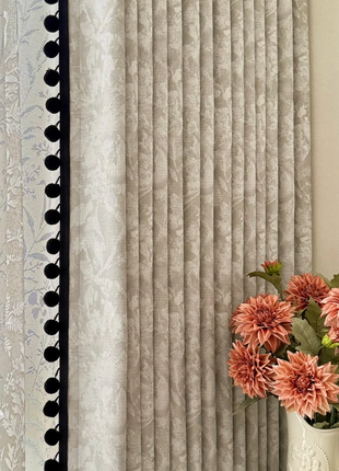 a white vase with pink flowers placed on a table, with a curtain featuring vertical lines and a black trim on the left side.