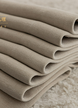 A close-up image of a stack of folded beige fabric, possibly a blanket or garment, with a text overlay in a foreign language.